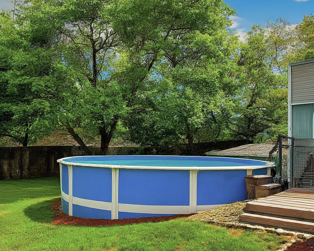 A blue above-ground pool in a backyard with trees, a deck, and a stone wall.